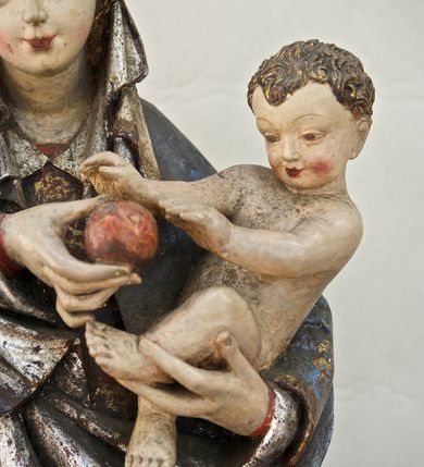 Zdjęcie nr 12: Rzeźba pełna, ukazująca Matkę Boską z Dzieciątkiem na niskiej, wielobocznej podstawie. Maria stoi, lewą ręką podtrzymuje Jezusa, a prawą podaje mu jabłko i spogląda na ten gest. Ma owalną, pełną twarz, o wysokim czole, wąskich brwiach, dużych, półprzymkniętych oczach, niewielkim nosie i ustach rozchylonych w delikatnym uśmiechu. Jest ubrana w czerwoną suknię ze złotym ornamentem, której fragment widać przy prawej nodze i bardzo obszerny, gęsto drapowany granatowy płaszcz ze złotym ornamentem i srebrnym podbiciem. Wierzchnie okrycie owija się wokół lewej ręki Madonny i szerokimi, twardo giętymi fałdami spływa w dół; przy prawym biodrze tworzy rurkowato zwinięte kaskady. Maria ma na głowę zarzucony welon, który, wywijając się, spływa na dekolt oraz wysoką, złotą, otwarta koronę. Rzeźba jest sygnowana z tyłu przy dolnej krawędzi „A(NDRZEJ) WALIJEWSKI”. Polichromia w partiach ciała naturalistyczna. 