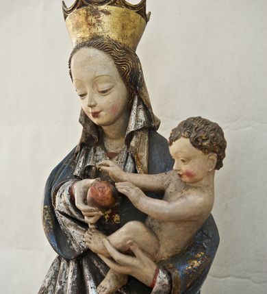 Zdjęcie nr 11: Rzeźba pełna, ukazująca Matkę Boską z Dzieciątkiem na niskiej, wielobocznej podstawie. Maria stoi, lewą ręką podtrzymuje Jezusa, a prawą podaje mu jabłko i spogląda na ten gest. Ma owalną, pełną twarz, o wysokim czole, wąskich brwiach, dużych, półprzymkniętych oczach, niewielkim nosie i ustach rozchylonych w delikatnym uśmiechu. Jest ubrana w czerwoną suknię ze złotym ornamentem, której fragment widać przy prawej nodze i bardzo obszerny, gęsto drapowany granatowy płaszcz ze złotym ornamentem i srebrnym podbiciem. Wierzchnie okrycie owija się wokół lewej ręki Madonny i szerokimi, twardo giętymi fałdami spływa w dół; przy prawym biodrze tworzy rurkowato zwinięte kaskady. Maria ma na głowę zarzucony welon, który, wywijając się, spływa na dekolt oraz wysoką, złotą, otwarta koronę. Rzeźba jest sygnowana z tyłu przy dolnej krawędzi „A(NDRZEJ) WALIJEWSKI”. Polichromia w partiach ciała naturalistyczna. 