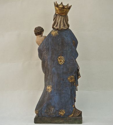 Zdjęcie nr 7: Rzeźba pełna, ukazująca Matkę Boską z Dzieciątkiem na niskiej, wielobocznej podstawie. Maria stoi, lewą ręką podtrzymuje Jezusa, a prawą podaje mu jabłko i spogląda na ten gest. Ma owalną, pełną twarz, o wysokim czole, wąskich brwiach, dużych, półprzymkniętych oczach, niewielkim nosie i ustach rozchylonych w delikatnym uśmiechu. Jest ubrana w czerwoną suknię ze złotym ornamentem, której fragment widać przy prawej nodze i bardzo obszerny, gęsto drapowany granatowy płaszcz ze złotym ornamentem i srebrnym podbiciem. Wierzchnie okrycie owija się wokół lewej ręki Madonny i szerokimi, twardo giętymi fałdami spływa w dół; przy prawym biodrze tworzy rurkowato zwinięte kaskady. Maria ma na głowę zarzucony welon, który, wywijając się, spływa na dekolt oraz wysoką, złotą, otwarta koronę. Rzeźba jest sygnowana z tyłu przy dolnej krawędzi „A(NDRZEJ) WALIJEWSKI”. Polichromia w partiach ciała naturalistyczna. 