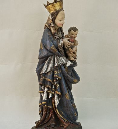 Zdjęcie nr 5: Rzeźba pełna, ukazująca Matkę Boską z Dzieciątkiem na niskiej, wielobocznej podstawie. Maria stoi, lewą ręką podtrzymuje Jezusa, a prawą podaje mu jabłko i spogląda na ten gest. Ma owalną, pełną twarz, o wysokim czole, wąskich brwiach, dużych, półprzymkniętych oczach, niewielkim nosie i ustach rozchylonych w delikatnym uśmiechu. Jest ubrana w czerwoną suknię ze złotym ornamentem, której fragment widać przy prawej nodze i bardzo obszerny, gęsto drapowany granatowy płaszcz ze złotym ornamentem i srebrnym podbiciem. Wierzchnie okrycie owija się wokół lewej ręki Madonny i szerokimi, twardo giętymi fałdami spływa w dół; przy prawym biodrze tworzy rurkowato zwinięte kaskady. Maria ma na głowę zarzucony welon, który, wywijając się, spływa na dekolt oraz wysoką, złotą, otwarta koronę. Rzeźba jest sygnowana z tyłu przy dolnej krawędzi „A(NDRZEJ) WALIJEWSKI”. Polichromia w partiach ciała naturalistyczna. 