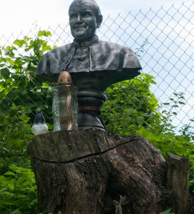 Zdjęcie nr 1: Popiersie Jana Pawła II na wysokim, profilowanym cokole. Papież ma owalną twarz, lekko uniesione brwi i zmarszczone czoło, wzrok skierowany w prawo, usta rozciągnięte w delikatnym uśmiechu. Widoczny jest fragment sutanny i pelerynki. Polichromia imituje odlew z metalu. 