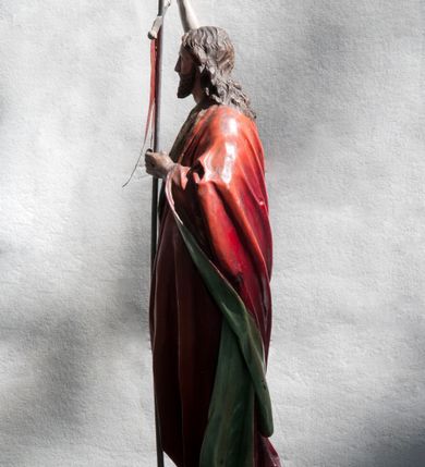 Zdjęcie nr 3: Rzeźba pełna ukazująca Chrystusa Zmartwychwstałego przedstawionego całopostaciowo, w lekkim kontrapoście, z delikatnie zaznaczoną muskulaturą i głową lekko opuszczoną na prawe ramię. Ma jasną karnację, twarz pociągłą o prostym nosie, okoloną długą brodą i falującymi brązowymi włosami spływającymi na plecy i ramiona. Na dłoniach i w boku widoczne ślady męki. Jezus jest ubrany w udrapowany, podbity zielenią czerwony płaszcz w diagonalnym układzie zarzucony na lewe ramię i owijający się wokół prawego biodra. W lewej dłoni trzyma chorągiew w postaci purpurowej tkaniny z krzyżem na drewnianym drzewcu, prawą ma uniesioną. Postać ustawiona na niewysokiej, ośmiobocznej podstawie.  