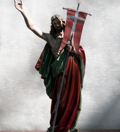 Zdjęcie nr 2: Rzeźba pełna ukazująca Chrystusa Zmartwychwstałego przedstawionego całopostaciowo, w lekkim kontrapoście, z delikatnie zaznaczoną muskulaturą i głową lekko opuszczoną na prawe ramię. Ma jasną karnację, twarz pociągłą o prostym nosie, okoloną długą brodą i falującymi brązowymi włosami spływającymi na plecy i ramiona. Na dłoniach i w boku widoczne ślady męki. Jezus jest ubrany w udrapowany, podbity zielenią czerwony płaszcz w diagonalnym układzie zarzucony na lewe ramię i owijający się wokół prawego biodra. W lewej dłoni trzyma chorągiew w postaci purpurowej tkaniny z krzyżem na drewnianym drzewcu, prawą ma uniesioną. Postać ustawiona na niewysokiej, ośmiobocznej podstawie.  