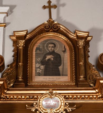 Zdjęcie nr 3: Ołtarz przyścienny, architektoniczny, jednokondygnacyjny ze zwieńczeniem, jednoosiowy. Mensa zdemontowana, pozostała część wsparta na korycnkich kolumnach. Retabulum flankowane kolumnami, na wysokich cokołach; trzony kolumn marmoryzowane, z nalożoną u dołu złoconą aplikacją z motywem kwiatowym. Na licach cokołów prostokątne płyciny wypełnione uskrzydlonymi główkami aniołków oraz motywem roślinnym. Kolumny podtrzymują przełamane, niepełne belkowanie. W polu ołtarza arkadowa nisza zakończona złoconą konchą; ujęta smukłymi, złoconymi kolumienkami kompozywowymi dźwigajacymi archiwoltę wypełnioną kimationem jońskim. Wewnątrz pełnoplastyczna, polichromowana figura św. Józefa. Nad niszą owalny kartusz z napisem „Święty Józefie / Módl się / za nami”. Belkowanie u góry dekorowane kimationem, powyżej przerwany przyczółek w formie wolut skierowanych do wewnątrz osi ołtarza. Zwieńczenie flankowane kolumnami oraz uszami w formie spływu wolutowego dekorowanego kampanulami i akantem, od góry zamknięte dwiema wolutami, zwieńczone krzyżem. W polu obraz św. Stanisława Kostki. Struktura malowana w kolorze jasnego brązu, kolumny marmoryzowane, ornamentyka i profilowania złocone.