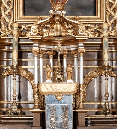 Zdjęcie nr 1: Tabernakulum dwudrzwiowe, w kształcie prostopadłościennej, obitej blachą metalową skrzynki. Na drzwiczkach odlewana dekoracja z kłosów i winnych gron, powyżej gorejące serce. Tabernakulum ujęte drewnianym tronem wystawienia w formie ażurowej kolumnady na planie półkola. Kolumny ustawione są na wysokich cokołach, wspierają niepełne belkowanie, zwieńczone sześcioma wolutami, spiętymi w kabłąk, tworzącymi formę połowy baldachimu. U szczytu korona zamknięta, pod nią Baranek Apokaliptyczny, a u spodu podwieszona szyszka otoczona liśćmi winogrona. Trzony kolumn w dolnej części zdobione są płaskorzeźbioną dekoracją roślinną, a w górnej części są gładkie, kapitele kompozytowe. Na tabernakulum ustawiona jest para adorujących aniołków, należących zapewne do kompletu tego zespołu. Aniołki ukazane są w pozycji klęczącej z rękami złożonymi w geście modlitwy i z pochylonymi głowami. Ubrane są w długie i złocone suknie.