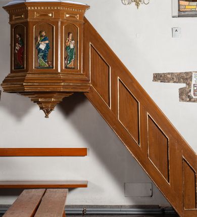 Zdjęcie nr 1: Ambona przyścienna, wisząca. Kosz ośmioboczny z silnie profilowaną podstawą, zakończony u dołu szyszką. Ścianki kosza zdobione pięcioma płycinami w formie stojących prostokątów, zamkniętych łukiem odcinkowym, z opaską malowaną na złoto. W każdej z czterech płycin płaskorzeźbione i polichromowane przedstawienie jednego z ewangelistów: Marka, Mateusza, Łukasza i Jana. Każda z postaci ukazana całopostaciowo, frontalnie, ze zwojem w lewej ręce, prawą czyniąca gest błogosławieństwa. U stóp ewangelistów przedstawiono odpowiadające im atrybuty: św. Marka – lew, św. Jana – orzeł, św. Mateusza – człowiek, św. Łukasza – wół. Balustrada ambony z belkowaniem, zdobionym motywem rautów we fryzie i kostkowaniem pod gzymsem. Ambona dostępna przez drewniane schody, od wschodu. Balustrada schodów zdobiona czterema romboidalnymi płycinami o brzegach malowanych na złoto. Struktura malowana w kolorze naturalnego drewna, detale i ornamentyka złocone.