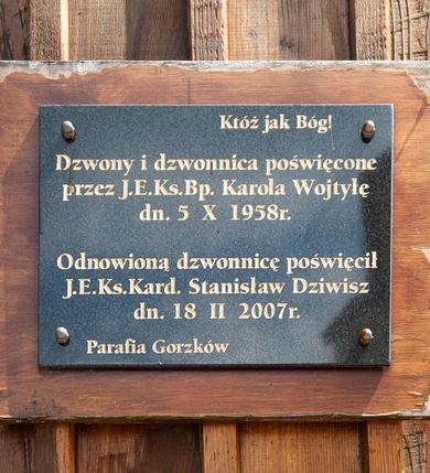 Zdjęcie nr 1: Tablica w kształcie leżącego prostokąta, umieszczona na drewnianej desce. W polu kuty, złocony napis „Któż jak Bóg! / Dzwony i dzwonnica poświęcone / przez J(ego) E(kscelencję) Ks(iędza) B(isku)p(a) Karola Wojtyłę / dn(ia) 5 X 1958 r(oku) / Odnowioną dzwonnicę poświęcił J(ego) E(minencja) Ks(iądz) Kard(ynał) Stanisław Dziwisz / dn(ia) 18 II 2007 r(oku) / Parafia Gorzków”.