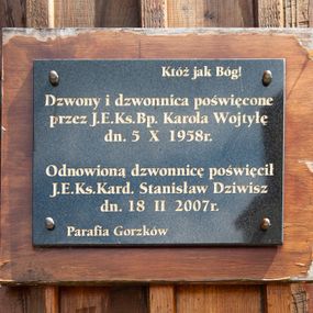 Zdjęcie nr 1: Tablica w kształcie leżącego prostokąta, umieszczona na drewnianej desce. W polu kuty, złocony napis „Któż jak Bóg! / Dzwony i dzwonnica poświęcone / przez J(ego) E(kscelencję) Ks(iędza) B(isku)p(a) Karola Wojtyłę / dn(ia) 5 X 1958 r(oku) / Odnowioną dzwonnicę poświęcił J(ego) E(minencja) Ks(iądz) Kard(ynał) Stanisław Dziwisz / dn(ia) 18 II 2007 r(oku) / Parafia Gorzków”.