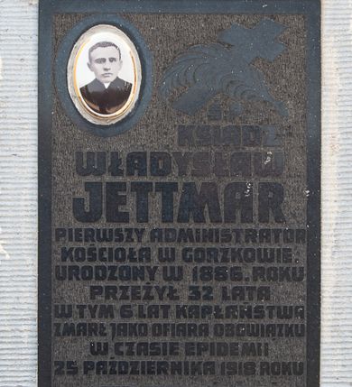 Zdjęcie nr 1: Tablica w kształcie stojącego prostokąta umieszczona na zewnętrznej ścianie południowej kaplicy pw. Michała Archanioła. W prawym, górnym narożu medalion ze zdjęciem, w lewym skrzyżowane liść palmy oraz krzyż. Poniżej napis  „Ś(WIĘTEJ) P(AMIĘCI) / KSIĄDZ / WŁADYSŁAW / JETTMAR / PIERWSZY ADMINISTRATOR / KOŚCIOŁA W GORZKOWIE / URODZONY W 1886. ROKU / PRZEŻYŁ 32 LATA / W TYM 6 LAT KAPŁAŃSTWA / ZMARŁ JAKO OFIARA OBOWIĄZKU / W CZASIE EPIDEMII / 25 PAŹDZIERNIKA 1918 ROKU / «NIKT NIE MOŻE MIEĆ / WIĘKSZEJ MIŁOŚCI / JAK ODDAĆ ŻYCIE / ZA SWYCH PRZYJACIÓŁ» / J.15.13. / PARAFIANIE 1967 R(OK)”. Tło fakturowane. Wokół tablicy cementowe obramienie  zdobione poziomym żłobkowaniem.