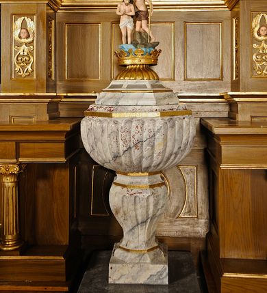 Zdjęcie nr 1: Chrzcielnica z pokrywą ustawiona na dwustopniowym, kamiennym cokole na rzucie kwadratu. Stopa w formie ośmiokąta foremnego na kształt jednolalkowej tralki, trzon żłobkowany. Czara ośmioboczna, puklowana. Pokrywa na ośmiobocznej, profilowanej podstawie z uszami po dwóch bokach, na bocznych i tylnych kwaterach napis „R(OKU) P(AŃSKIEGO) 1923 FUNDAT RZEPA z BUGAJU”.  Zwieńczona złoconą koroną, od dołu ujętą puklowaniem, na której ustawiona rzeźbiona, pełnoplastyczna scena Chrztu Chrystusa w Jordanie. Chrystus ukazany ze złożonymi rękami, stoi w wodzie, powyżej na skałach Jan Chrzciciel trzymający w lewej ręce wysoki krzyż, a w prawej konchę. Nad nimi wydłużona gloria z gołębicą Ducha Świętego. Tło sceny stanowi drzewo.