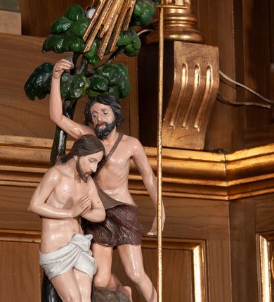 Zdjęcie nr 4: Chrzcielnica z pokrywą ustawiona na dwustopniowym, kamiennym cokole na rzucie kwadratu. Stopa w formie ośmiokąta foremnego na kształt jednolalkowej tralki, trzon żłobkowany. Czara ośmioboczna, puklowana. Pokrywa na ośmiobocznej, profilowanej podstawie z uszami po dwóch bokach, na bocznych i tylnych kwaterach napis „R(OKU) P(AŃSKIEGO) 1923 FUNDAT RZEPA z BUGAJU”.  Zwieńczona złoconą koroną, od dołu ujętą puklowaniem, na której ustawiona rzeźbiona, pełnoplastyczna scena Chrztu Chrystusa w Jordanie. Chrystus ukazany ze złożonymi rękami, stoi w wodzie, powyżej na skałach Jan Chrzciciel trzymający w lewej ręce wysoki krzyż, a w prawej konchę. Nad nimi wydłużona gloria z gołębicą Ducha Świętego. Tło sceny stanowi drzewo.