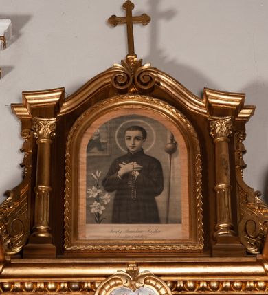 Zdjęcie nr 1: Obraz w kształcie stojącego prostokąta zamkniętego półkoliście, umieszczony w złoconej, profilowanej ramie, przeszklony. Przedstawia św. Stanisława Kostkę. Święty ukazany frontalnie, w półpostaci, z rękami skrzyżowanymi na piersi, z różańcem w dłoniach. Ubrany jest w czarną sutannę jezuicką. Głowa otoczona okrągłym nimbem. Po lewej stronie obrazu gałązka białej lilii, wyżej fragment obrazu z Matką Boską z Dzieciątkiem, a po prawej bukłak z wodą na wysokim drzewcu. W dole obrazu inskrypcja „Święty Stanisław Kostka / Patron młodzieży”.