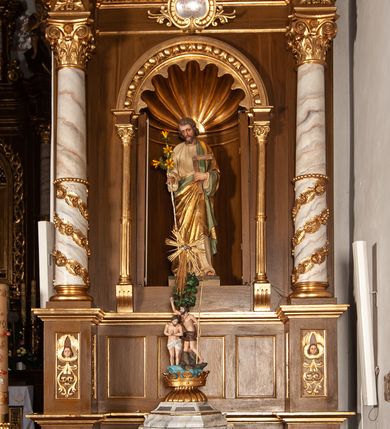 Zdjęcie nr 2: Rzeźba św. Józefa w niszy lewego ołtarza bocznego św. Józefa. Figura umieszczona na niewielkim, prostopadłościennym cokole. Święty przedstawiony w postawie stojącej z lekko ugiętym lewym kolanem. Ubrany w długą  i białą tunikę przewiązaną w pasie, na lewe ramię narzucony złoty płaszcz z zieloną podszewką, który oplata jego biodra. Twarz podłużna z przymkniętymi oczami, długim i prostym nosem, wąskimi ustami. Włosy i zarost – brązowe. W prawej ręce trzyma żółtą lilię, w lewej węgielnicę.