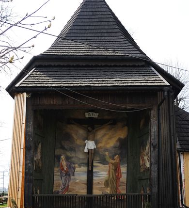 Zdjęcie nr 2: Na zewnętrznej stronie prezbiterium znajduje się ogrójec, który zamknięty jest trzema ścianami, otwarty od frontu i przegrodzony balustradą, zamknięty od góry zadaszeniem. Na północnej i południowej ścianie znajdują się po trzy pola w malowanych prześwitach, dwa dolne wypełniają sceny męki Chrystusa, w najwyższych przedstawiono zachmurzone niebo. Scena główna znajduje się na wschodniej ścianie prezbiterium i złożona jest z rzeźbionego krucyfiksu oraz malowanego tła. Cykl przedstawień męki Pańskiej rozpoczyna się na ścianie południowej. Scena pierwsza cyklu – Modlitwa w Ogrójcu, usytuowana jest w środkowym polu ściany. Ilustruje klęczącego na tle pejzażu przed skałą Chrystusa, przed którym w promieniach światła unosi się anioł trzymający kielich. W prawej części przedstawienia, w tle ukazani są dwaj śpiący apostołowie. Scena druga, Biczowanie, usytuowana jest w najniższym polu ściany południowej, przedstawia wnętrze z kamiennymi ścianami. W jej prawej części znajduje się na wpół leżący i oparty o słup Chrystus ze związanymi rękami, przepasany czerwoną szatą z koroną cierniową na głowie. W lewej części przedstawiony jest oprawca, ukazany tyłem, opierający się lewą nogą o ciało Jezusa i zamachujący się na niego biczem. Kolejne przedstawienia z cyklu znajdują się na północnej ścianie. Kontynuuje go trzecia scena – Koronowanie cierniem, znajdująca się w najniższym polu. Ukazuje wnętrze z niebieską kotarą w tle, w lewej części przedstawiony jest siedzący frontalnie Chrystus w czerwonym płaszczu, za nim zaś mężczyzna, nakładający na jego głowę koronę cierniową. W prawej części na pierwszym planie znajdują się mężczyźni naigrywający się z Jezusa i składający mu pokłon, z tyłu postać żołnierza. Scena czwarta to Droga Krzyżowa, usytuowana jest w środkowym polu ściany północnej. Ilustruje upadek Chrystusa niosącego krzyż i Szymona Cyrenejczyka, który pomaga mu go podnieść. W tle pejzaż z tłumem ludzi i żołnierzami. Scena główna przedstawia Ukrzyżowanie z Marią i św. Janem. W centrum znajduje się krzyż z figurą umarłego Chrystusa na tle malowanego pustynno-górzystego pejzażu z Jerozolimą oraz ze skłębionym żółto-błękitnym niebem. Po lewej stronie krzyża Matka Boska zwrócona w trzech czwartych w prawo, lewą ręką wskazuje na krzyż, prawą składa na piersi. Ubrana jest w czerwoną suknię i niebieski płaszcz narzucony na głowę. Po prawej stronie krzyża św. Jan ukazany z profilu, prawą nogą oparty o skałę, wznosi ku górze splecione dłonie w geście rozpaczy. Ubrany w zieloną suknię, przepasaną w talii oraz czerwony płaszcz. Wnętrze kapliczki pomalowane jest na kolor jasnozielony. 


