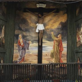 Zdjęcie nr 1: Na zewnętrznej stronie prezbiterium znajduje się ogrójec, który zamknięty jest trzema ścianami, otwarty od frontu i przegrodzony balustradą, zamknięty od góry zadaszeniem. Na północnej i południowej ścianie znajdują się po trzy pola w malowanych prześwitach, dwa dolne wypełniają sceny męki Chrystusa, w najwyższych przedstawiono zachmurzone niebo. Scena główna znajduje się na wschodniej ścianie prezbiterium i złożona jest z rzeźbionego krucyfiksu oraz malowanego tła. Cykl przedstawień męki Pańskiej rozpoczyna się na ścianie południowej. Scena pierwsza cyklu – Modlitwa w Ogrójcu, usytuowana jest w środkowym polu ściany. Ilustruje klęczącego na tle pejzażu przed skałą Chrystusa, przed którym w promieniach światła unosi się anioł trzymający kielich. W prawej części przedstawienia, w tle ukazani są dwaj śpiący apostołowie. Scena druga, Biczowanie, usytuowana jest w najniższym polu ściany południowej, przedstawia wnętrze z kamiennymi ścianami. W jej prawej części znajduje się na wpół leżący i oparty o słup Chrystus ze związanymi rękami, przepasany czerwoną szatą z koroną cierniową na głowie. W lewej części przedstawiony jest oprawca, ukazany tyłem, opierający się lewą nogą o ciało Jezusa i zamachujący się na niego biczem. Kolejne przedstawienia z cyklu znajdują się na północnej ścianie. Kontynuuje go trzecia scena – Koronowanie cierniem, znajdująca się w najniższym polu. Ukazuje wnętrze z niebieską kotarą w tle, w lewej części przedstawiony jest siedzący frontalnie Chrystus w czerwonym płaszczu, za nim zaś mężczyzna, nakładający na jego głowę koronę cierniową. W prawej części na pierwszym planie znajdują się mężczyźni naigrywający się z Jezusa i składający mu pokłon, z tyłu postać żołnierza. Scena czwarta to Droga Krzyżowa, usytuowana jest w środkowym polu ściany północnej. Ilustruje upadek Chrystusa niosącego krzyż i Szymona Cyrenejczyka, który pomaga mu go podnieść. W tle pejzaż z tłumem ludzi i żołnierzami. Scena główna przedstawia Ukrzyżowanie z Marią i św. Janem. W centrum znajduje się krzyż z figurą umarłego Chrystusa na tle malowanego pustynno-górzystego pejzażu z Jerozolimą oraz ze skłębionym żółto-błękitnym niebem. Po lewej stronie krzyża Matka Boska zwrócona w trzech czwartych w prawo, lewą ręką wskazuje na krzyż, prawą składa na piersi. Ubrana jest w czerwoną suknię i niebieski płaszcz narzucony na głowę. Po prawej stronie krzyża św. Jan ukazany z profilu, prawą nogą oparty o skałę, wznosi ku górze splecione dłonie w geście rozpaczy. Ubrany w zieloną suknię, przepasaną w talii oraz czerwony płaszcz. Wnętrze kapliczki pomalowane jest na kolor jasnozielony. 


