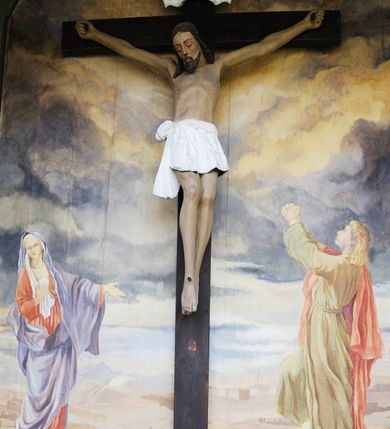 Zdjęcie nr 13: Na zewnętrznej stronie prezbiterium znajduje się ogrójec, który zamknięty jest trzema ścianami, otwarty od frontu i przegrodzony balustradą, zamknięty od góry zadaszeniem. Na północnej i południowej ścianie znajdują się po trzy pola w malowanych prześwitach, dwa dolne wypełniają sceny męki Chrystusa, w najwyższych przedstawiono zachmurzone niebo. Scena główna znajduje się na wschodniej ścianie prezbiterium i złożona jest z rzeźbionego krucyfiksu oraz malowanego tła. Cykl przedstawień męki Pańskiej rozpoczyna się na ścianie południowej. Scena pierwsza cyklu – Modlitwa w Ogrójcu, usytuowana jest w środkowym polu ściany. Ilustruje klęczącego na tle pejzażu przed skałą Chrystusa, przed którym w promieniach światła unosi się anioł trzymający kielich. W prawej części przedstawienia, w tle ukazani są dwaj śpiący apostołowie. Scena druga, Biczowanie, usytuowana jest w najniższym polu ściany południowej, przedstawia wnętrze z kamiennymi ścianami. W jej prawej części znajduje się na wpół leżący i oparty o słup Chrystus ze związanymi rękami, przepasany czerwoną szatą z koroną cierniową na głowie. W lewej części przedstawiony jest oprawca, ukazany tyłem, opierający się lewą nogą o ciało Jezusa i zamachujący się na niego biczem. Kolejne przedstawienia z cyklu znajdują się na północnej ścianie. Kontynuuje go trzecia scena – Koronowanie cierniem, znajdująca się w najniższym polu. Ukazuje wnętrze z niebieską kotarą w tle, w lewej części przedstawiony jest siedzący frontalnie Chrystus w czerwonym płaszczu, za nim zaś mężczyzna, nakładający na jego głowę koronę cierniową. W prawej części na pierwszym planie znajdują się mężczyźni naigrywający się z Jezusa i składający mu pokłon, z tyłu postać żołnierza. Scena czwarta to Droga Krzyżowa, usytuowana jest w środkowym polu ściany północnej. Ilustruje upadek Chrystusa niosącego krzyż i Szymona Cyrenejczyka, który pomaga mu go podnieść. W tle pejzaż z tłumem ludzi i żołnierzami. Scena główna przedstawia Ukrzyżowanie z Marią i św. Janem. W centrum znajduje się krzyż z figurą umarłego Chrystusa na tle malowanego pustynno-górzystego pejzażu z Jerozolimą oraz ze skłębionym żółto-błękitnym niebem. Po lewej stronie krzyża Matka Boska zwrócona w trzech czwartych w prawo, lewą ręką wskazuje na krzyż, prawą składa na piersi. Ubrana jest w czerwoną suknię i niebieski płaszcz narzucony na głowę. Po prawej stronie krzyża św. Jan ukazany z profilu, prawą nogą oparty o skałę, wznosi ku górze splecione dłonie w geście rozpaczy. Ubrany w zieloną suknię, przepasaną w talii oraz czerwony płaszcz. Wnętrze kapliczki pomalowane jest na kolor jasnozielony. 


