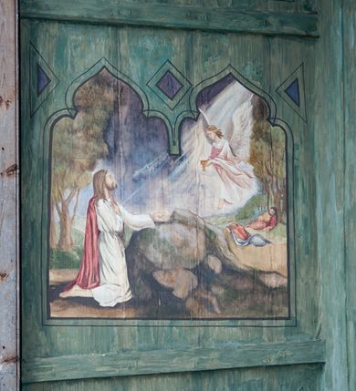 Zdjęcie nr 17: Na zewnętrznej stronie prezbiterium znajduje się ogrójec, który zamknięty jest trzema ścianami, otwarty od frontu i przegrodzony balustradą, zamknięty od góry zadaszeniem. Na północnej i południowej ścianie znajdują się po trzy pola w malowanych prześwitach, dwa dolne wypełniają sceny męki Chrystusa, w najwyższych przedstawiono zachmurzone niebo. Scena główna znajduje się na wschodniej ścianie prezbiterium i złożona jest z rzeźbionego krucyfiksu oraz malowanego tła. Cykl przedstawień męki Pańskiej rozpoczyna się na ścianie południowej. Scena pierwsza cyklu – Modlitwa w Ogrójcu, usytuowana jest w środkowym polu ściany. Ilustruje klęczącego na tle pejzażu przed skałą Chrystusa, przed którym w promieniach światła unosi się anioł trzymający kielich. W prawej części przedstawienia, w tle ukazani są dwaj śpiący apostołowie. Scena druga, Biczowanie, usytuowana jest w najniższym polu ściany południowej, przedstawia wnętrze z kamiennymi ścianami. W jej prawej części znajduje się na wpół leżący i oparty o słup Chrystus ze związanymi rękami, przepasany czerwoną szatą z koroną cierniową na głowie. W lewej części przedstawiony jest oprawca, ukazany tyłem, opierający się lewą nogą o ciało Jezusa i zamachujący się na niego biczem. Kolejne przedstawienia z cyklu znajdują się na północnej ścianie. Kontynuuje go trzecia scena – Koronowanie cierniem, znajdująca się w najniższym polu. Ukazuje wnętrze z niebieską kotarą w tle, w lewej części przedstawiony jest siedzący frontalnie Chrystus w czerwonym płaszczu, za nim zaś mężczyzna, nakładający na jego głowę koronę cierniową. W prawej części na pierwszym planie znajdują się mężczyźni naigrywający się z Jezusa i składający mu pokłon, z tyłu postać żołnierza. Scena czwarta to Droga Krzyżowa, usytuowana jest w środkowym polu ściany północnej. Ilustruje upadek Chrystusa niosącego krzyż i Szymona Cyrenejczyka, który pomaga mu go podnieść. W tle pejzaż z tłumem ludzi i żołnierzami. Scena główna przedstawia Ukrzyżowanie z Marią i św. Janem. W centrum znajduje się krzyż z figurą umarłego Chrystusa na tle malowanego pustynno-górzystego pejzażu z Jerozolimą oraz ze skłębionym żółto-błękitnym niebem. Po lewej stronie krzyża Matka Boska zwrócona w trzech czwartych w prawo, lewą ręką wskazuje na krzyż, prawą składa na piersi. Ubrana jest w czerwoną suknię i niebieski płaszcz narzucony na głowę. Po prawej stronie krzyża św. Jan ukazany z profilu, prawą nogą oparty o skałę, wznosi ku górze splecione dłonie w geście rozpaczy. Ubrany w zieloną suknię, przepasaną w talii oraz czerwony płaszcz. Wnętrze kapliczki pomalowane jest na kolor jasnozielony. 


