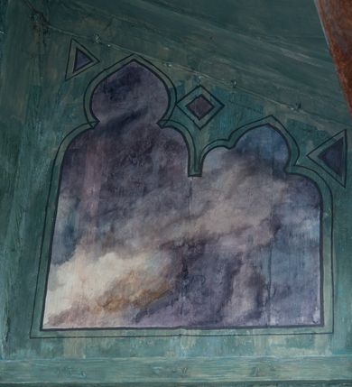 Zdjęcie nr 11: Na zewnętrznej stronie prezbiterium znajduje się ogrójec, który zamknięty jest trzema ścianami, otwarty od frontu i przegrodzony balustradą, zamknięty od góry zadaszeniem. Na północnej i południowej ścianie znajdują się po trzy pola w malowanych prześwitach, dwa dolne wypełniają sceny męki Chrystusa, w najwyższych przedstawiono zachmurzone niebo. Scena główna znajduje się na wschodniej ścianie prezbiterium i złożona jest z rzeźbionego krucyfiksu oraz malowanego tła. Cykl przedstawień męki Pańskiej rozpoczyna się na ścianie południowej. Scena pierwsza cyklu – Modlitwa w Ogrójcu, usytuowana jest w środkowym polu ściany. Ilustruje klęczącego na tle pejzażu przed skałą Chrystusa, przed którym w promieniach światła unosi się anioł trzymający kielich. W prawej części przedstawienia, w tle ukazani są dwaj śpiący apostołowie. Scena druga, Biczowanie, usytuowana jest w najniższym polu ściany południowej, przedstawia wnętrze z kamiennymi ścianami. W jej prawej części znajduje się na wpół leżący i oparty o słup Chrystus ze związanymi rękami, przepasany czerwoną szatą z koroną cierniową na głowie. W lewej części przedstawiony jest oprawca, ukazany tyłem, opierający się lewą nogą o ciało Jezusa i zamachujący się na niego biczem. Kolejne przedstawienia z cyklu znajdują się na północnej ścianie. Kontynuuje go trzecia scena – Koronowanie cierniem, znajdująca się w najniższym polu. Ukazuje wnętrze z niebieską kotarą w tle, w lewej części przedstawiony jest siedzący frontalnie Chrystus w czerwonym płaszczu, za nim zaś mężczyzna, nakładający na jego głowę koronę cierniową. W prawej części na pierwszym planie znajdują się mężczyźni naigrywający się z Jezusa i składający mu pokłon, z tyłu postać żołnierza. Scena czwarta to Droga Krzyżowa, usytuowana jest w środkowym polu ściany północnej. Ilustruje upadek Chrystusa niosącego krzyż i Szymona Cyrenejczyka, który pomaga mu go podnieść. W tle pejzaż z tłumem ludzi i żołnierzami. Scena główna przedstawia Ukrzyżowanie z Marią i św. Janem. W centrum znajduje się krzyż z figurą umarłego Chrystusa na tle malowanego pustynno-górzystego pejzażu z Jerozolimą oraz ze skłębionym żółto-błękitnym niebem. Po lewej stronie krzyża Matka Boska zwrócona w trzech czwartych w prawo, lewą ręką wskazuje na krzyż, prawą składa na piersi. Ubrana jest w czerwoną suknię i niebieski płaszcz narzucony na głowę. Po prawej stronie krzyża św. Jan ukazany z profilu, prawą nogą oparty o skałę, wznosi ku górze splecione dłonie w geście rozpaczy. Ubrany w zieloną suknię, przepasaną w talii oraz czerwony płaszcz. Wnętrze kapliczki pomalowane jest na kolor jasnozielony. 


