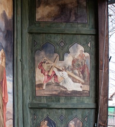 Zdjęcie nr 6: Na zewnętrznej stronie prezbiterium znajduje się ogrójec, który zamknięty jest trzema ścianami, otwarty od frontu i przegrodzony balustradą, zamknięty od góry zadaszeniem. Na północnej i południowej ścianie znajdują się po trzy pola w malowanych prześwitach, dwa dolne wypełniają sceny męki Chrystusa, w najwyższych przedstawiono zachmurzone niebo. Scena główna znajduje się na wschodniej ścianie prezbiterium i złożona jest z rzeźbionego krucyfiksu oraz malowanego tła. Cykl przedstawień męki Pańskiej rozpoczyna się na ścianie południowej. Scena pierwsza cyklu – Modlitwa w Ogrójcu, usytuowana jest w środkowym polu ściany. Ilustruje klęczącego na tle pejzażu przed skałą Chrystusa, przed którym w promieniach światła unosi się anioł trzymający kielich. W prawej części przedstawienia, w tle ukazani są dwaj śpiący apostołowie. Scena druga, Biczowanie, usytuowana jest w najniższym polu ściany południowej, przedstawia wnętrze z kamiennymi ścianami. W jej prawej części znajduje się na wpół leżący i oparty o słup Chrystus ze związanymi rękami, przepasany czerwoną szatą z koroną cierniową na głowie. W lewej części przedstawiony jest oprawca, ukazany tyłem, opierający się lewą nogą o ciało Jezusa i zamachujący się na niego biczem. Kolejne przedstawienia z cyklu znajdują się na północnej ścianie. Kontynuuje go trzecia scena – Koronowanie cierniem, znajdująca się w najniższym polu. Ukazuje wnętrze z niebieską kotarą w tle, w lewej części przedstawiony jest siedzący frontalnie Chrystus w czerwonym płaszczu, za nim zaś mężczyzna, nakładający na jego głowę koronę cierniową. W prawej części na pierwszym planie znajdują się mężczyźni naigrywający się z Jezusa i składający mu pokłon, z tyłu postać żołnierza. Scena czwarta to Droga Krzyżowa, usytuowana jest w środkowym polu ściany północnej. Ilustruje upadek Chrystusa niosącego krzyż i Szymona Cyrenejczyka, który pomaga mu go podnieść. W tle pejzaż z tłumem ludzi i żołnierzami. Scena główna przedstawia Ukrzyżowanie z Marią i św. Janem. W centrum znajduje się krzyż z figurą umarłego Chrystusa na tle malowanego pustynno-górzystego pejzażu z Jerozolimą oraz ze skłębionym żółto-błękitnym niebem. Po lewej stronie krzyża Matka Boska zwrócona w trzech czwartych w prawo, lewą ręką wskazuje na krzyż, prawą składa na piersi. Ubrana jest w czerwoną suknię i niebieski płaszcz narzucony na głowę. Po prawej stronie krzyża św. Jan ukazany z profilu, prawą nogą oparty o skałę, wznosi ku górze splecione dłonie w geście rozpaczy. Ubrany w zieloną suknię, przepasaną w talii oraz czerwony płaszcz. Wnętrze kapliczki pomalowane jest na kolor jasnozielony. 


