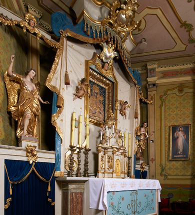 Zdjęcie nr 7: Ołtarz prostopadłościenny, murowany, w drewnianej obudowie, z drewnianą mensą. Antependium w kształcie leżącego prostokąta ujętego profilowaną ramą, dekorowane monogramem imienia MARIA w środku pola i symetrycznie rozłożonymi po bokach różanymi gałązkami. Na mensie ustawione architektoniczne tabernakulum. 
Nastawa w formie paludamentu podwiązanego po bokach sznurem z chwostami, lamowanego frędzlą, zwieńczonego baldachimem utworzonym z fragmentu wklęsło-wypukłego gzymsu z lambrekinem i rokokowym kartuszem. W polu paludamentu wnęka w kształcie stojącego prostokąta o wklęsłych górnych narożach w profilowanej ramie, ustawiona na cokole ujętym spływami wolutowymi. Wewnątrz obraz Matki Boskiej z Dzieciątkiem ujęty grubą ramą dekorowaną akantem i palmetami, ponad nim uskrzydlona główka anielska. Wnękę flankują dwa unoszące się putta, powyżej niej kartusz z monogramem Marii. Paludament dekorowany plastycznymi gwiazdami. Po bokach nastawy dwie bramki z otworami wejściowymi w formie stojącego prostokąta, zamkniętymi prostym gzymsem ze znajdującymi się powyżej postumentami dekorowanymi różami, na których stoją dwie figury aniołów. W górnej części, z obydwu stron nastawy poziome listwy konstrukcyjne mocujące całość retabulum do ścian bocznych prezbiterium, dekorowane są uskrzydlonymi główkami anielskimi z muszlami oraz roślinnymi festonami. Paludament polichromowany wewnątrz na biało, zewnątrz na niebiesko z pozłacanymi detalami.

