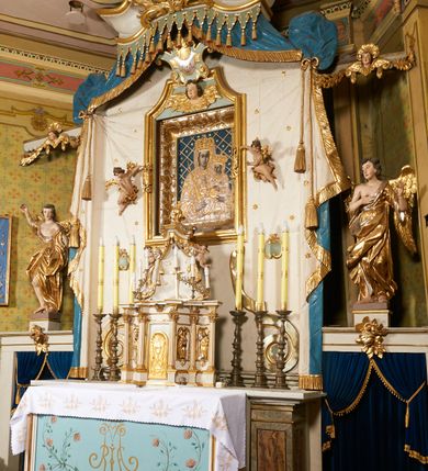 Zdjęcie nr 5: Ołtarz prostopadłościenny, murowany, w drewnianej obudowie, z drewnianą mensą. Antependium w kształcie leżącego prostokąta ujętego profilowaną ramą, dekorowane monogramem imienia MARIA w środku pola i symetrycznie rozłożonymi po bokach różanymi gałązkami. Na mensie ustawione architektoniczne tabernakulum. 
Nastawa w formie paludamentu podwiązanego po bokach sznurem z chwostami, lamowanego frędzlą, zwieńczonego baldachimem utworzonym z fragmentu wklęsło-wypukłego gzymsu z lambrekinem i rokokowym kartuszem. W polu paludamentu wnęka w kształcie stojącego prostokąta o wklęsłych górnych narożach w profilowanej ramie, ustawiona na cokole ujętym spływami wolutowymi. Wewnątrz obraz Matki Boskiej z Dzieciątkiem ujęty grubą ramą dekorowaną akantem i palmetami, ponad nim uskrzydlona główka anielska. Wnękę flankują dwa unoszące się putta, powyżej niej kartusz z monogramem Marii. Paludament dekorowany plastycznymi gwiazdami. Po bokach nastawy dwie bramki z otworami wejściowymi w formie stojącego prostokąta, zamkniętymi prostym gzymsem ze znajdującymi się powyżej postumentami dekorowanymi różami, na których stoją dwie figury aniołów. W górnej części, z obydwu stron nastawy poziome listwy konstrukcyjne mocujące całość retabulum do ścian bocznych prezbiterium, dekorowane są uskrzydlonymi główkami anielskimi z muszlami oraz roślinnymi festonami. Paludament polichromowany wewnątrz na biało, zewnątrz na niebiesko z pozłacanymi detalami.

