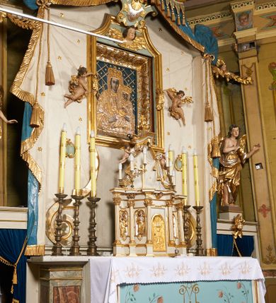 Zdjęcie nr 9: Ołtarz prostopadłościenny, murowany, w drewnianej obudowie, z drewnianą mensą. Antependium w kształcie leżącego prostokąta ujętego profilowaną ramą, dekorowane monogramem imienia MARIA w środku pola i symetrycznie rozłożonymi po bokach różanymi gałązkami. Na mensie ustawione architektoniczne tabernakulum. 
Nastawa w formie paludamentu podwiązanego po bokach sznurem z chwostami, lamowanego frędzlą, zwieńczonego baldachimem utworzonym z fragmentu wklęsło-wypukłego gzymsu z lambrekinem i rokokowym kartuszem. W polu paludamentu wnęka w kształcie stojącego prostokąta o wklęsłych górnych narożach w profilowanej ramie, ustawiona na cokole ujętym spływami wolutowymi. Wewnątrz obraz Matki Boskiej z Dzieciątkiem ujęty grubą ramą dekorowaną akantem i palmetami, ponad nim uskrzydlona główka anielska. Wnękę flankują dwa unoszące się putta, powyżej niej kartusz z monogramem Marii. Paludament dekorowany plastycznymi gwiazdami. Po bokach nastawy dwie bramki z otworami wejściowymi w formie stojącego prostokąta, zamkniętymi prostym gzymsem ze znajdującymi się powyżej postumentami dekorowanymi różami, na których stoją dwie figury aniołów. W górnej części, z obydwu stron nastawy poziome listwy konstrukcyjne mocujące całość retabulum do ścian bocznych prezbiterium, dekorowane są uskrzydlonymi główkami anielskimi z muszlami oraz roślinnymi festonami. Paludament polichromowany wewnątrz na biało, zewnątrz na niebiesko z pozłacanymi detalami.

