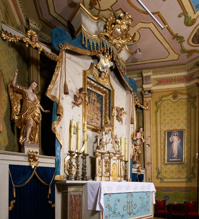 Zdjęcie nr 6: Ołtarz prostopadłościenny, murowany, w drewnianej obudowie, z drewnianą mensą. Antependium w kształcie leżącego prostokąta ujętego profilowaną ramą, dekorowane monogramem imienia MARIA w środku pola i symetrycznie rozłożonymi po bokach różanymi gałązkami. Na mensie ustawione architektoniczne tabernakulum. 
Nastawa w formie paludamentu podwiązanego po bokach sznurem z chwostami, lamowanego frędzlą, zwieńczonego baldachimem utworzonym z fragmentu wklęsło-wypukłego gzymsu z lambrekinem i rokokowym kartuszem. W polu paludamentu wnęka w kształcie stojącego prostokąta o wklęsłych górnych narożach w profilowanej ramie, ustawiona na cokole ujętym spływami wolutowymi. Wewnątrz obraz Matki Boskiej z Dzieciątkiem ujęty grubą ramą dekorowaną akantem i palmetami, ponad nim uskrzydlona główka anielska. Wnękę flankują dwa unoszące się putta, powyżej niej kartusz z monogramem Marii. Paludament dekorowany plastycznymi gwiazdami. Po bokach nastawy dwie bramki z otworami wejściowymi w formie stojącego prostokąta, zamkniętymi prostym gzymsem ze znajdującymi się powyżej postumentami dekorowanymi różami, na których stoją dwie figury aniołów. W górnej części, z obydwu stron nastawy poziome listwy konstrukcyjne mocujące całość retabulum do ścian bocznych prezbiterium, dekorowane są uskrzydlonymi główkami anielskimi z muszlami oraz roślinnymi festonami. Paludament polichromowany wewnątrz na biało, zewnątrz na niebiesko z pozłacanymi detalami.

