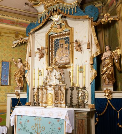 Zdjęcie nr 4: Ołtarz prostopadłościenny, murowany, w drewnianej obudowie, z drewnianą mensą. Antependium w kształcie leżącego prostokąta ujętego profilowaną ramą, dekorowane monogramem imienia MARIA w środku pola i symetrycznie rozłożonymi po bokach różanymi gałązkami. Na mensie ustawione architektoniczne tabernakulum. 
Nastawa w formie paludamentu podwiązanego po bokach sznurem z chwostami, lamowanego frędzlą, zwieńczonego baldachimem utworzonym z fragmentu wklęsło-wypukłego gzymsu z lambrekinem i rokokowym kartuszem. W polu paludamentu wnęka w kształcie stojącego prostokąta o wklęsłych górnych narożach w profilowanej ramie, ustawiona na cokole ujętym spływami wolutowymi. Wewnątrz obraz Matki Boskiej z Dzieciątkiem ujęty grubą ramą dekorowaną akantem i palmetami, ponad nim uskrzydlona główka anielska. Wnękę flankują dwa unoszące się putta, powyżej niej kartusz z monogramem Marii. Paludament dekorowany plastycznymi gwiazdami. Po bokach nastawy dwie bramki z otworami wejściowymi w formie stojącego prostokąta, zamkniętymi prostym gzymsem ze znajdującymi się powyżej postumentami dekorowanymi różami, na których stoją dwie figury aniołów. W górnej części, z obydwu stron nastawy poziome listwy konstrukcyjne mocujące całość retabulum do ścian bocznych prezbiterium, dekorowane są uskrzydlonymi główkami anielskimi z muszlami oraz roślinnymi festonami. Paludament polichromowany wewnątrz na biało, zewnątrz na niebiesko z pozłacanymi detalami.

