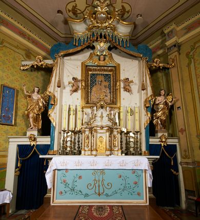 Zdjęcie nr 2: Ołtarz prostopadłościenny, murowany, w drewnianej obudowie, z drewnianą mensą. Antependium w kształcie leżącego prostokąta ujętego profilowaną ramą, dekorowane monogramem imienia MARIA w środku pola i symetrycznie rozłożonymi po bokach różanymi gałązkami. Na mensie ustawione architektoniczne tabernakulum. 
Nastawa w formie paludamentu podwiązanego po bokach sznurem z chwostami, lamowanego frędzlą, zwieńczonego baldachimem utworzonym z fragmentu wklęsło-wypukłego gzymsu z lambrekinem i rokokowym kartuszem. W polu paludamentu wnęka w kształcie stojącego prostokąta o wklęsłych górnych narożach w profilowanej ramie, ustawiona na cokole ujętym spływami wolutowymi. Wewnątrz obraz Matki Boskiej z Dzieciątkiem ujęty grubą ramą dekorowaną akantem i palmetami, ponad nim uskrzydlona główka anielska. Wnękę flankują dwa unoszące się putta, powyżej niej kartusz z monogramem Marii. Paludament dekorowany plastycznymi gwiazdami. Po bokach nastawy dwie bramki z otworami wejściowymi w formie stojącego prostokąta, zamkniętymi prostym gzymsem ze znajdującymi się powyżej postumentami dekorowanymi różami, na których stoją dwie figury aniołów. W górnej części, z obydwu stron nastawy poziome listwy konstrukcyjne mocujące całość retabulum do ścian bocznych prezbiterium, dekorowane są uskrzydlonymi główkami anielskimi z muszlami oraz roślinnymi festonami. Paludament polichromowany wewnątrz na biało, zewnątrz na niebiesko z pozłacanymi detalami.

