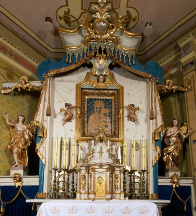 Zdjęcie nr 1: Ołtarz prostopadłościenny, murowany, w drewnianej obudowie, z drewnianą mensą. Antependium w kształcie leżącego prostokąta ujętego profilowaną ramą, dekorowane monogramem imienia MARIA w środku pola i symetrycznie rozłożonymi po bokach różanymi gałązkami. Na mensie ustawione architektoniczne tabernakulum. 
Nastawa w formie paludamentu podwiązanego po bokach sznurem z chwostami, lamowanego frędzlą, zwieńczonego baldachimem utworzonym z fragmentu wklęsło-wypukłego gzymsu z lambrekinem i rokokowym kartuszem. W polu paludamentu wnęka w kształcie stojącego prostokąta o wklęsłych górnych narożach w profilowanej ramie, ustawiona na cokole ujętym spływami wolutowymi. Wewnątrz obraz Matki Boskiej z Dzieciątkiem ujęty grubą ramą dekorowaną akantem i palmetami, ponad nim uskrzydlona główka anielska. Wnękę flankują dwa unoszące się putta, powyżej niej kartusz z monogramem Marii. Paludament dekorowany plastycznymi gwiazdami. Po bokach nastawy dwie bramki z otworami wejściowymi w formie stojącego prostokąta, zamkniętymi prostym gzymsem ze znajdującymi się powyżej postumentami dekorowanymi różami, na których stoją dwie figury aniołów. W górnej części, z obydwu stron nastawy poziome listwy konstrukcyjne mocujące całość retabulum do ścian bocznych prezbiterium, dekorowane są uskrzydlonymi główkami anielskimi z muszlami oraz roślinnymi festonami. Paludament polichromowany wewnątrz na biało, zewnątrz na niebiesko z pozłacanymi detalami.

