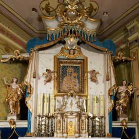 Zdjęcie nr 1: Ołtarz prostopadłościenny, murowany, w drewnianej obudowie, z drewnianą mensą. Antependium w kształcie leżącego prostokąta ujętego profilowaną ramą, dekorowane monogramem imienia MARIA w środku pola i symetrycznie rozłożonymi po bokach różanymi gałązkami. Na mensie ustawione architektoniczne tabernakulum. 
Nastawa w formie paludamentu podwiązanego po bokach sznurem z chwostami, lamowanego frędzlą, zwieńczonego baldachimem utworzonym z fragmentu wklęsło-wypukłego gzymsu z lambrekinem i rokokowym kartuszem. W polu paludamentu wnęka w kształcie stojącego prostokąta o wklęsłych górnych narożach w profilowanej ramie, ustawiona na cokole ujętym spływami wolutowymi. Wewnątrz obraz Matki Boskiej z Dzieciątkiem ujęty grubą ramą dekorowaną akantem i palmetami, ponad nim uskrzydlona główka anielska. Wnękę flankują dwa unoszące się putta, powyżej niej kartusz z monogramem Marii. Paludament dekorowany plastycznymi gwiazdami. Po bokach nastawy dwie bramki z otworami wejściowymi w formie stojącego prostokąta, zamkniętymi prostym gzymsem ze znajdującymi się powyżej postumentami dekorowanymi różami, na których stoją dwie figury aniołów. W górnej części, z obydwu stron nastawy poziome listwy konstrukcyjne mocujące całość retabulum do ścian bocznych prezbiterium, dekorowane są uskrzydlonymi główkami anielskimi z muszlami oraz roślinnymi festonami. Paludament polichromowany wewnątrz na biało, zewnątrz na niebiesko z pozłacanymi detalami.

