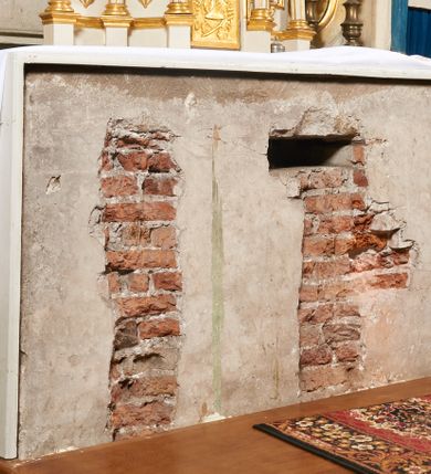 Zdjęcie nr 104: Ołtarz prostopadłościenny, murowany, w drewnianej obudowie, z drewnianą mensą. Antependium w kształcie leżącego prostokąta ujętego profilowaną ramą, dekorowane monogramem imienia MARIA w środku pola i symetrycznie rozłożonymi po bokach różanymi gałązkami. Na mensie ustawione architektoniczne tabernakulum. 
Nastawa w formie paludamentu podwiązanego po bokach sznurem z chwostami, lamowanego frędzlą, zwieńczonego baldachimem utworzonym z fragmentu wklęsło-wypukłego gzymsu z lambrekinem i rokokowym kartuszem. W polu paludamentu wnęka w kształcie stojącego prostokąta o wklęsłych górnych narożach w profilowanej ramie, ustawiona na cokole ujętym spływami wolutowymi. Wewnątrz obraz Matki Boskiej z Dzieciątkiem ujęty grubą ramą dekorowaną akantem i palmetami, ponad nim uskrzydlona główka anielska. Wnękę flankują dwa unoszące się putta, powyżej niej kartusz z monogramem Marii. Paludament dekorowany plastycznymi gwiazdami. Po bokach nastawy dwie bramki z otworami wejściowymi w formie stojącego prostokąta, zamkniętymi prostym gzymsem ze znajdującymi się powyżej postumentami dekorowanymi różami, na których stoją dwie figury aniołów. W górnej części, z obydwu stron nastawy poziome listwy konstrukcyjne mocujące całość retabulum do ścian bocznych prezbiterium, dekorowane są uskrzydlonymi główkami anielskimi z muszlami oraz roślinnymi festonami. Paludament polichromowany wewnątrz na biało, zewnątrz na niebiesko z pozłacanymi detalami.

