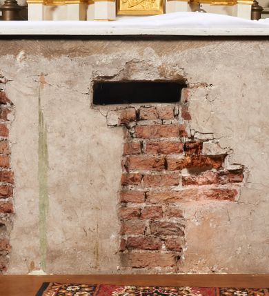 Zdjęcie nr 105: Ołtarz prostopadłościenny, murowany, w drewnianej obudowie, z drewnianą mensą. Antependium w kształcie leżącego prostokąta ujętego profilowaną ramą, dekorowane monogramem imienia MARIA w środku pola i symetrycznie rozłożonymi po bokach różanymi gałązkami. Na mensie ustawione architektoniczne tabernakulum. 
Nastawa w formie paludamentu podwiązanego po bokach sznurem z chwostami, lamowanego frędzlą, zwieńczonego baldachimem utworzonym z fragmentu wklęsło-wypukłego gzymsu z lambrekinem i rokokowym kartuszem. W polu paludamentu wnęka w kształcie stojącego prostokąta o wklęsłych górnych narożach w profilowanej ramie, ustawiona na cokole ujętym spływami wolutowymi. Wewnątrz obraz Matki Boskiej z Dzieciątkiem ujęty grubą ramą dekorowaną akantem i palmetami, ponad nim uskrzydlona główka anielska. Wnękę flankują dwa unoszące się putta, powyżej niej kartusz z monogramem Marii. Paludament dekorowany plastycznymi gwiazdami. Po bokach nastawy dwie bramki z otworami wejściowymi w formie stojącego prostokąta, zamkniętymi prostym gzymsem ze znajdującymi się powyżej postumentami dekorowanymi różami, na których stoją dwie figury aniołów. W górnej części, z obydwu stron nastawy poziome listwy konstrukcyjne mocujące całość retabulum do ścian bocznych prezbiterium, dekorowane są uskrzydlonymi główkami anielskimi z muszlami oraz roślinnymi festonami. Paludament polichromowany wewnątrz na biało, zewnątrz na niebiesko z pozłacanymi detalami.

