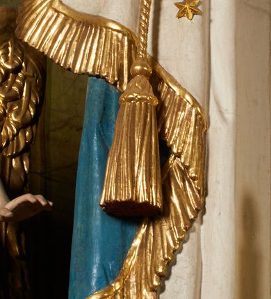 Zdjęcie nr 34: Ołtarz prostopadłościenny, murowany, w drewnianej obudowie, z drewnianą mensą. Antependium w kształcie leżącego prostokąta ujętego profilowaną ramą, dekorowane monogramem imienia MARIA w środku pola i symetrycznie rozłożonymi po bokach różanymi gałązkami. Na mensie ustawione architektoniczne tabernakulum. 
Nastawa w formie paludamentu podwiązanego po bokach sznurem z chwostami, lamowanego frędzlą, zwieńczonego baldachimem utworzonym z fragmentu wklęsło-wypukłego gzymsu z lambrekinem i rokokowym kartuszem. W polu paludamentu wnęka w kształcie stojącego prostokąta o wklęsłych górnych narożach w profilowanej ramie, ustawiona na cokole ujętym spływami wolutowymi. Wewnątrz obraz Matki Boskiej z Dzieciątkiem ujęty grubą ramą dekorowaną akantem i palmetami, ponad nim uskrzydlona główka anielska. Wnękę flankują dwa unoszące się putta, powyżej niej kartusz z monogramem Marii. Paludament dekorowany plastycznymi gwiazdami. Po bokach nastawy dwie bramki z otworami wejściowymi w formie stojącego prostokąta, zamkniętymi prostym gzymsem ze znajdującymi się powyżej postumentami dekorowanymi różami, na których stoją dwie figury aniołów. W górnej części, z obydwu stron nastawy poziome listwy konstrukcyjne mocujące całość retabulum do ścian bocznych prezbiterium, dekorowane są uskrzydlonymi główkami anielskimi z muszlami oraz roślinnymi festonami. Paludament polichromowany wewnątrz na biało, zewnątrz na niebiesko z pozłacanymi detalami.

