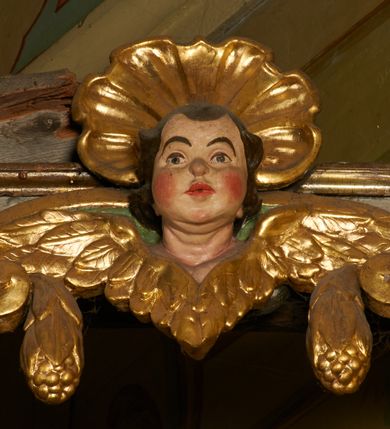 Zdjęcie nr 28: Ołtarz prostopadłościenny, murowany, w drewnianej obudowie, z drewnianą mensą. Antependium w kształcie leżącego prostokąta ujętego profilowaną ramą, dekorowane monogramem imienia MARIA w środku pola i symetrycznie rozłożonymi po bokach różanymi gałązkami. Na mensie ustawione architektoniczne tabernakulum. 
Nastawa w formie paludamentu podwiązanego po bokach sznurem z chwostami, lamowanego frędzlą, zwieńczonego baldachimem utworzonym z fragmentu wklęsło-wypukłego gzymsu z lambrekinem i rokokowym kartuszem. W polu paludamentu wnęka w kształcie stojącego prostokąta o wklęsłych górnych narożach w profilowanej ramie, ustawiona na cokole ujętym spływami wolutowymi. Wewnątrz obraz Matki Boskiej z Dzieciątkiem ujęty grubą ramą dekorowaną akantem i palmetami, ponad nim uskrzydlona główka anielska. Wnękę flankują dwa unoszące się putta, powyżej niej kartusz z monogramem Marii. Paludament dekorowany plastycznymi gwiazdami. Po bokach nastawy dwie bramki z otworami wejściowymi w formie stojącego prostokąta, zamkniętymi prostym gzymsem ze znajdującymi się powyżej postumentami dekorowanymi różami, na których stoją dwie figury aniołów. W górnej części, z obydwu stron nastawy poziome listwy konstrukcyjne mocujące całość retabulum do ścian bocznych prezbiterium, dekorowane są uskrzydlonymi główkami anielskimi z muszlami oraz roślinnymi festonami. Paludament polichromowany wewnątrz na biało, zewnątrz na niebiesko z pozłacanymi detalami.

