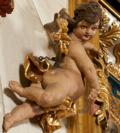 Zdjęcie nr 97: Ołtarz prostopadłościenny, murowany, w drewnianej obudowie, z drewnianą mensą. Antependium w kształcie leżącego prostokąta ujętego profilowaną ramą, dekorowane monogramem imienia MARIA w środku pola i symetrycznie rozłożonymi po bokach różanymi gałązkami. Na mensie ustawione architektoniczne tabernakulum. 
Nastawa w formie paludamentu podwiązanego po bokach sznurem z chwostami, lamowanego frędzlą, zwieńczonego baldachimem utworzonym z fragmentu wklęsło-wypukłego gzymsu z lambrekinem i rokokowym kartuszem. W polu paludamentu wnęka w kształcie stojącego prostokąta o wklęsłych górnych narożach w profilowanej ramie, ustawiona na cokole ujętym spływami wolutowymi. Wewnątrz obraz Matki Boskiej z Dzieciątkiem ujęty grubą ramą dekorowaną akantem i palmetami, ponad nim uskrzydlona główka anielska. Wnękę flankują dwa unoszące się putta, powyżej niej kartusz z monogramem Marii. Paludament dekorowany plastycznymi gwiazdami. Po bokach nastawy dwie bramki z otworami wejściowymi w formie stojącego prostokąta, zamkniętymi prostym gzymsem ze znajdującymi się powyżej postumentami dekorowanymi różami, na których stoją dwie figury aniołów. W górnej części, z obydwu stron nastawy poziome listwy konstrukcyjne mocujące całość retabulum do ścian bocznych prezbiterium, dekorowane są uskrzydlonymi główkami anielskimi z muszlami oraz roślinnymi festonami. Paludament polichromowany wewnątrz na biało, zewnątrz na niebiesko z pozłacanymi detalami.

