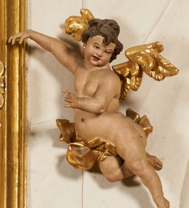 Zdjęcie nr 81: Ołtarz prostopadłościenny, murowany, w drewnianej obudowie, z drewnianą mensą. Antependium w kształcie leżącego prostokąta ujętego profilowaną ramą, dekorowane monogramem imienia MARIA w środku pola i symetrycznie rozłożonymi po bokach różanymi gałązkami. Na mensie ustawione architektoniczne tabernakulum. 
Nastawa w formie paludamentu podwiązanego po bokach sznurem z chwostami, lamowanego frędzlą, zwieńczonego baldachimem utworzonym z fragmentu wklęsło-wypukłego gzymsu z lambrekinem i rokokowym kartuszem. W polu paludamentu wnęka w kształcie stojącego prostokąta o wklęsłych górnych narożach w profilowanej ramie, ustawiona na cokole ujętym spływami wolutowymi. Wewnątrz obraz Matki Boskiej z Dzieciątkiem ujęty grubą ramą dekorowaną akantem i palmetami, ponad nim uskrzydlona główka anielska. Wnękę flankują dwa unoszące się putta, powyżej niej kartusz z monogramem Marii. Paludament dekorowany plastycznymi gwiazdami. Po bokach nastawy dwie bramki z otworami wejściowymi w formie stojącego prostokąta, zamkniętymi prostym gzymsem ze znajdującymi się powyżej postumentami dekorowanymi różami, na których stoją dwie figury aniołów. W górnej części, z obydwu stron nastawy poziome listwy konstrukcyjne mocujące całość retabulum do ścian bocznych prezbiterium, dekorowane są uskrzydlonymi główkami anielskimi z muszlami oraz roślinnymi festonami. Paludament polichromowany wewnątrz na biało, zewnątrz na niebiesko z pozłacanymi detalami.

