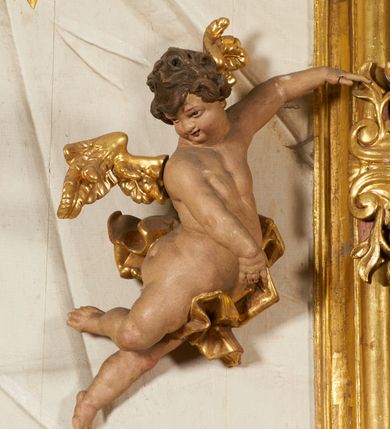 Zdjęcie nr 95: Ołtarz prostopadłościenny, murowany, w drewnianej obudowie, z drewnianą mensą. Antependium w kształcie leżącego prostokąta ujętego profilowaną ramą, dekorowane monogramem imienia MARIA w środku pola i symetrycznie rozłożonymi po bokach różanymi gałązkami. Na mensie ustawione architektoniczne tabernakulum. 
Nastawa w formie paludamentu podwiązanego po bokach sznurem z chwostami, lamowanego frędzlą, zwieńczonego baldachimem utworzonym z fragmentu wklęsło-wypukłego gzymsu z lambrekinem i rokokowym kartuszem. W polu paludamentu wnęka w kształcie stojącego prostokąta o wklęsłych górnych narożach w profilowanej ramie, ustawiona na cokole ujętym spływami wolutowymi. Wewnątrz obraz Matki Boskiej z Dzieciątkiem ujęty grubą ramą dekorowaną akantem i palmetami, ponad nim uskrzydlona główka anielska. Wnękę flankują dwa unoszące się putta, powyżej niej kartusz z monogramem Marii. Paludament dekorowany plastycznymi gwiazdami. Po bokach nastawy dwie bramki z otworami wejściowymi w formie stojącego prostokąta, zamkniętymi prostym gzymsem ze znajdującymi się powyżej postumentami dekorowanymi różami, na których stoją dwie figury aniołów. W górnej części, z obydwu stron nastawy poziome listwy konstrukcyjne mocujące całość retabulum do ścian bocznych prezbiterium, dekorowane są uskrzydlonymi główkami anielskimi z muszlami oraz roślinnymi festonami. Paludament polichromowany wewnątrz na biało, zewnątrz na niebiesko z pozłacanymi detalami.

