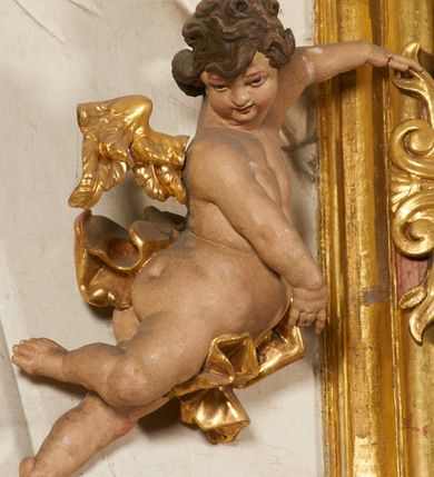 Zdjęcie nr 96: Ołtarz prostopadłościenny, murowany, w drewnianej obudowie, z drewnianą mensą. Antependium w kształcie leżącego prostokąta ujętego profilowaną ramą, dekorowane monogramem imienia MARIA w środku pola i symetrycznie rozłożonymi po bokach różanymi gałązkami. Na mensie ustawione architektoniczne tabernakulum. 
Nastawa w formie paludamentu podwiązanego po bokach sznurem z chwostami, lamowanego frędzlą, zwieńczonego baldachimem utworzonym z fragmentu wklęsło-wypukłego gzymsu z lambrekinem i rokokowym kartuszem. W polu paludamentu wnęka w kształcie stojącego prostokąta o wklęsłych górnych narożach w profilowanej ramie, ustawiona na cokole ujętym spływami wolutowymi. Wewnątrz obraz Matki Boskiej z Dzieciątkiem ujęty grubą ramą dekorowaną akantem i palmetami, ponad nim uskrzydlona główka anielska. Wnękę flankują dwa unoszące się putta, powyżej niej kartusz z monogramem Marii. Paludament dekorowany plastycznymi gwiazdami. Po bokach nastawy dwie bramki z otworami wejściowymi w formie stojącego prostokąta, zamkniętymi prostym gzymsem ze znajdującymi się powyżej postumentami dekorowanymi różami, na których stoją dwie figury aniołów. W górnej części, z obydwu stron nastawy poziome listwy konstrukcyjne mocujące całość retabulum do ścian bocznych prezbiterium, dekorowane są uskrzydlonymi główkami anielskimi z muszlami oraz roślinnymi festonami. Paludament polichromowany wewnątrz na biało, zewnątrz na niebiesko z pozłacanymi detalami.

