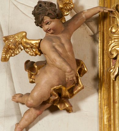 Zdjęcie nr 94: Ołtarz prostopadłościenny, murowany, w drewnianej obudowie, z drewnianą mensą. Antependium w kształcie leżącego prostokąta ujętego profilowaną ramą, dekorowane monogramem imienia MARIA w środku pola i symetrycznie rozłożonymi po bokach różanymi gałązkami. Na mensie ustawione architektoniczne tabernakulum. 
Nastawa w formie paludamentu podwiązanego po bokach sznurem z chwostami, lamowanego frędzlą, zwieńczonego baldachimem utworzonym z fragmentu wklęsło-wypukłego gzymsu z lambrekinem i rokokowym kartuszem. W polu paludamentu wnęka w kształcie stojącego prostokąta o wklęsłych górnych narożach w profilowanej ramie, ustawiona na cokole ujętym spływami wolutowymi. Wewnątrz obraz Matki Boskiej z Dzieciątkiem ujęty grubą ramą dekorowaną akantem i palmetami, ponad nim uskrzydlona główka anielska. Wnękę flankują dwa unoszące się putta, powyżej niej kartusz z monogramem Marii. Paludament dekorowany plastycznymi gwiazdami. Po bokach nastawy dwie bramki z otworami wejściowymi w formie stojącego prostokąta, zamkniętymi prostym gzymsem ze znajdującymi się powyżej postumentami dekorowanymi różami, na których stoją dwie figury aniołów. W górnej części, z obydwu stron nastawy poziome listwy konstrukcyjne mocujące całość retabulum do ścian bocznych prezbiterium, dekorowane są uskrzydlonymi główkami anielskimi z muszlami oraz roślinnymi festonami. Paludament polichromowany wewnątrz na biało, zewnątrz na niebiesko z pozłacanymi detalami.

