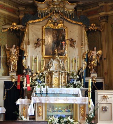 Zdjęcie nr 109: Ołtarz prostopadłościenny, murowany, w drewnianej obudowie, z drewnianą mensą. Antependium w kształcie leżącego prostokąta ujętego profilowaną ramą, dekorowane monogramem imienia MARIA w środku pola i symetrycznie rozłożonymi po bokach różanymi gałązkami. Na mensie ustawione architektoniczne tabernakulum. 
Nastawa w formie paludamentu podwiązanego po bokach sznurem z chwostami, lamowanego frędzlą, zwieńczonego baldachimem utworzonym z fragmentu wklęsło-wypukłego gzymsu z lambrekinem i rokokowym kartuszem. W polu paludamentu wnęka w kształcie stojącego prostokąta o wklęsłych górnych narożach w profilowanej ramie, ustawiona na cokole ujętym spływami wolutowymi. Wewnątrz obraz Matki Boskiej z Dzieciątkiem ujęty grubą ramą dekorowaną akantem i palmetami, ponad nim uskrzydlona główka anielska. Wnękę flankują dwa unoszące się putta, powyżej niej kartusz z monogramem Marii. Paludament dekorowany plastycznymi gwiazdami. Po bokach nastawy dwie bramki z otworami wejściowymi w formie stojącego prostokąta, zamkniętymi prostym gzymsem ze znajdującymi się powyżej postumentami dekorowanymi różami, na których stoją dwie figury aniołów. W górnej części, z obydwu stron nastawy poziome listwy konstrukcyjne mocujące całość retabulum do ścian bocznych prezbiterium, dekorowane są uskrzydlonymi główkami anielskimi z muszlami oraz roślinnymi festonami. Paludament polichromowany wewnątrz na biało, zewnątrz na niebiesko z pozłacanymi detalami.

