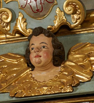 Zdjęcie nr 4: Obraz w kształcie pionowego prostokąta, z przedstawieniem Matki Boskiej z Dzieciątkiem, na niebieskim tle dekorowanym srebrzoną kratką, z malowanymi na drewnie partiami twarzy, dłoni i stóp oraz partiami szat i insygniami zasłoniętymi przez sukienki. Matka Boska ukazana w półpostaci, zwrócona delikatnie w trzech czwartych w lewą stronę, na lewym przedramieniu podtrzymuje Dzieciątko, a w prawej dłoni trzyma berło. Twarz pociągła, owalna, o dużych oczach, prostym, długim nosie i pełnych ustach, z odsłoniętym dużym prawym uchem. Łagodne rysy wydobyte zostały ostrym kontrastem światłocienia. Na głowie ma koronę zamkniętą, wokół głowy glorię promienistą i nimb zamknięty. Ubrana jest w suknię, dekorowaną gałązkami kwiatowymi, przepasaną w talii wstążką oraz płaszcz narzucony na ramiona, z ośmioramienną gwiazdą na prawym ramieniu, dekorowany ornamentem rocaille'owym z ośmioramienną gwiazdą na prawym ramieniu. Dzieciątko w pozycji siedzącej, zwrócone jest w trzech czwartych w prawą stronę, prawą dłonią błogosławi, lewą przytrzymuje na kolanie jabłko królewskie. Jego twarz jest okrągła o rysach wydobytych silnym światłocieniem, o dużych oczach, niewielkim nosie i pełnych ustach z odsłoniętym lewym, dużym uchem. Na głowie ma koronę zamkniętą, wokół twarzy glorię promienistą oraz nimb zamknięty. Ubrane jest w tunikę dekorowaną gałązkami kwiatowymi i lamówką. Obraz ujęty posrebrzaną ramą z plastycznym, rokokowym i pozłacanym ornamentem. 