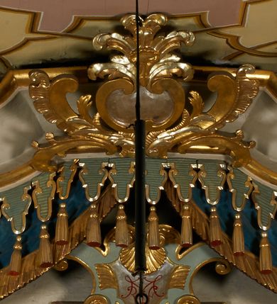 Zdjęcie nr 9: Obraz w kształcie pionowego prostokąta, z przedstawieniem Matki Boskiej z Dzieciątkiem, na niebieskim tle dekorowanym srebrzoną kratką, z malowanymi na drewnie partiami twarzy, dłoni i stóp oraz partiami szat i insygniami zasłoniętymi przez sukienki. Matka Boska ukazana w półpostaci, zwrócona delikatnie w trzech czwartych w lewą stronę, na lewym przedramieniu podtrzymuje Dzieciątko, a w prawej dłoni trzyma berło. Twarz pociągła, owalna, o dużych oczach, prostym, długim nosie i pełnych ustach, z odsłoniętym dużym prawym uchem. Łagodne rysy wydobyte zostały ostrym kontrastem światłocienia. Na głowie ma koronę zamkniętą, wokół głowy glorię promienistą i nimb zamknięty. Ubrana jest w suknię, dekorowaną gałązkami kwiatowymi, przepasaną w talii wstążką oraz płaszcz narzucony na ramiona, z ośmioramienną gwiazdą na prawym ramieniu, dekorowany ornamentem rocaille&#039;owym z ośmioramienną gwiazdą na prawym ramieniu. Dzieciątko w pozycji siedzącej, zwrócone jest w trzech czwartych w prawą stronę, prawą dłonią błogosławi, lewą przytrzymuje na kolanie jabłko królewskie. Jego twarz jest okrągła o rysach wydobytych silnym światłocieniem, o dużych oczach, niewielkim nosie i pełnych ustach z odsłoniętym lewym, dużym uchem. Na głowie ma koronę zamkniętą, wokół twarzy glorię promienistą oraz nimb zamknięty. Ubrane jest w tunikę dekorowaną gałązkami kwiatowymi i lamówką. Obraz ujęty posrebrzaną ramą z plastycznym, rokokowym i pozłacanym ornamentem. 