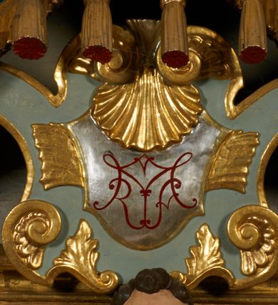 Zdjęcie nr 12: Obraz w kształcie pionowego prostokąta, z przedstawieniem Matki Boskiej z Dzieciątkiem, na niebieskim tle dekorowanym srebrzoną kratką, z malowanymi na drewnie partiami twarzy, dłoni i stóp oraz partiami szat i insygniami zasłoniętymi przez sukienki. Matka Boska ukazana w półpostaci, zwrócona delikatnie w trzech czwartych w lewą stronę, na lewym przedramieniu podtrzymuje Dzieciątko, a w prawej dłoni trzyma berło. Twarz pociągła, owalna, o dużych oczach, prostym, długim nosie i pełnych ustach, z odsłoniętym dużym prawym uchem. Łagodne rysy wydobyte zostały ostrym kontrastem światłocienia. Na głowie ma koronę zamkniętą, wokół głowy glorię promienistą i nimb zamknięty. Ubrana jest w suknię, dekorowaną gałązkami kwiatowymi, przepasaną w talii wstążką oraz płaszcz narzucony na ramiona, z ośmioramienną gwiazdą na prawym ramieniu, dekorowany ornamentem rocaille&#039;owym z ośmioramienną gwiazdą na prawym ramieniu. Dzieciątko w pozycji siedzącej, zwrócone jest w trzech czwartych w prawą stronę, prawą dłonią błogosławi, lewą przytrzymuje na kolanie jabłko królewskie. Jego twarz jest okrągła o rysach wydobytych silnym światłocieniem, o dużych oczach, niewielkim nosie i pełnych ustach z odsłoniętym lewym, dużym uchem. Na głowie ma koronę zamkniętą, wokół twarzy glorię promienistą oraz nimb zamknięty. Ubrane jest w tunikę dekorowaną gałązkami kwiatowymi i lamówką. Obraz ujęty posrebrzaną ramą z plastycznym, rokokowym i pozłacanym ornamentem. 