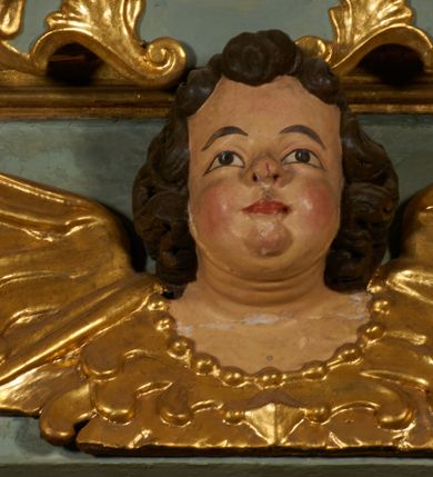 Zdjęcie nr 5: Obraz w kształcie pionowego prostokąta, z przedstawieniem Matki Boskiej z Dzieciątkiem, na niebieskim tle dekorowanym srebrzoną kratką, z malowanymi na drewnie partiami twarzy, dłoni i stóp oraz partiami szat i insygniami zasłoniętymi przez sukienki. Matka Boska ukazana w półpostaci, zwrócona delikatnie w trzech czwartych w lewą stronę, na lewym przedramieniu podtrzymuje Dzieciątko, a w prawej dłoni trzyma berło. Twarz pociągła, owalna, o dużych oczach, prostym, długim nosie i pełnych ustach, z odsłoniętym dużym prawym uchem. Łagodne rysy wydobyte zostały ostrym kontrastem światłocienia. Na głowie ma koronę zamkniętą, wokół głowy glorię promienistą i nimb zamknięty. Ubrana jest w suknię, dekorowaną gałązkami kwiatowymi, przepasaną w talii wstążką oraz płaszcz narzucony na ramiona, z ośmioramienną gwiazdą na prawym ramieniu, dekorowany ornamentem rocaille&#039;owym z ośmioramienną gwiazdą na prawym ramieniu. Dzieciątko w pozycji siedzącej, zwrócone jest w trzech czwartych w prawą stronę, prawą dłonią błogosławi, lewą przytrzymuje na kolanie jabłko królewskie. Jego twarz jest okrągła o rysach wydobytych silnym światłocieniem, o dużych oczach, niewielkim nosie i pełnych ustach z odsłoniętym lewym, dużym uchem. Na głowie ma koronę zamkniętą, wokół twarzy glorię promienistą oraz nimb zamknięty. Ubrane jest w tunikę dekorowaną gałązkami kwiatowymi i lamówką. Obraz ujęty posrebrzaną ramą z plastycznym, rokokowym i pozłacanym ornamentem. 