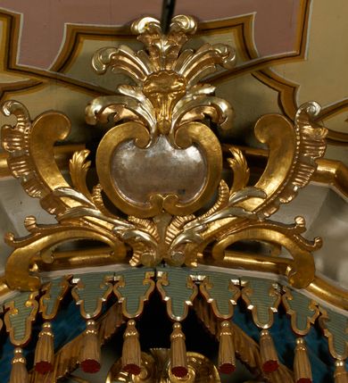 Zdjęcie nr 11: Obraz w kształcie pionowego prostokąta, z przedstawieniem Matki Boskiej z Dzieciątkiem, na niebieskim tle dekorowanym srebrzoną kratką, z malowanymi na drewnie partiami twarzy, dłoni i stóp oraz partiami szat i insygniami zasłoniętymi przez sukienki. Matka Boska ukazana w półpostaci, zwrócona delikatnie w trzech czwartych w lewą stronę, na lewym przedramieniu podtrzymuje Dzieciątko, a w prawej dłoni trzyma berło. Twarz pociągła, owalna, o dużych oczach, prostym, długim nosie i pełnych ustach, z odsłoniętym dużym prawym uchem. Łagodne rysy wydobyte zostały ostrym kontrastem światłocienia. Na głowie ma koronę zamkniętą, wokół głowy glorię promienistą i nimb zamknięty. Ubrana jest w suknię, dekorowaną gałązkami kwiatowymi, przepasaną w talii wstążką oraz płaszcz narzucony na ramiona, z ośmioramienną gwiazdą na prawym ramieniu, dekorowany ornamentem rocaille'owym z ośmioramienną gwiazdą na prawym ramieniu. Dzieciątko w pozycji siedzącej, zwrócone jest w trzech czwartych w prawą stronę, prawą dłonią błogosławi, lewą przytrzymuje na kolanie jabłko królewskie. Jego twarz jest okrągła o rysach wydobytych silnym światłocieniem, o dużych oczach, niewielkim nosie i pełnych ustach z odsłoniętym lewym, dużym uchem. Na głowie ma koronę zamkniętą, wokół twarzy glorię promienistą oraz nimb zamknięty. Ubrane jest w tunikę dekorowaną gałązkami kwiatowymi i lamówką. Obraz ujęty posrebrzaną ramą z plastycznym, rokokowym i pozłacanym ornamentem. 