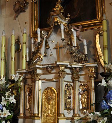 Zdjęcie nr 2: Tabernakulum architektoniczne, jednokondygnacyjne, pięcioosiowe. Osie wydzielone są sześcioma kanelowanymi kolumienkami na wysokich, profilowanych cokołach, z liściastymi kapitelami, podtrzymującymi profilowane belkowanie wyłamane na ich osiach. W polach pomiędzy kolumnami znajdują się nisze ujęte profilowaną ramą z uszakami. W czterech bocznych niszach, na trójdzielnych, profilowanych wspornikach stoją płaskorzeźbione postacie czterech ewangelistów na tle muszli palmetowych. Figurki są całopostaciowe, w ujęciu frontalnym, w złotych szatach, podtrzymują otwarte księgi, wskazując na tekst. Drzwiczki znajdują się na osi środkowej, zamknięte są półkoliście, płaskorzeźbione z przedstawieniem kielicha z koroną cierniową i z hostią w glorii promienistej. Kielich flankowany jest kłosami zbóż i winną latoroślą oraz winogronami. W partii belkowania płaskorzeźbiona dekoracja złożona z naprzemiennie ułożonych główek anielskich o złotych skrzydłach i płaskich rozetek. Tron wystawienia w formie baldachimu, z lambrekinem i chwostami, zwieńczony jest kulą z krzyżem, z podwieszonym z tyłu paludamentem podtrzymywanym przez parę puttów. Struktura polichromowana na biało, profile i detale pozłocone. 