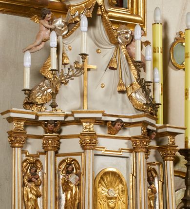 Zdjęcie nr 4: Tabernakulum architektoniczne, jednokondygnacyjne, pięcioosiowe. Osie wydzielone są sześcioma kanelowanymi kolumienkami na wysokich, profilowanych cokołach, z liściastymi kapitelami, podtrzymującymi profilowane belkowanie wyłamane na ich osiach. W polach pomiędzy kolumnami znajdują się nisze ujęte profilowaną ramą z uszakami. W czterech bocznych niszach, na trójdzielnych, profilowanych wspornikach stoją płaskorzeźbione postacie czterech ewangelistów na tle muszli palmetowych. Figurki są całopostaciowe, w ujęciu frontalnym, w złotych szatach, podtrzymują otwarte księgi, wskazując na tekst. Drzwiczki znajdują się na osi środkowej, zamknięte są półkoliście, płaskorzeźbione z przedstawieniem kielicha z koroną cierniową i z hostią w glorii promienistej. Kielich flankowany jest kłosami zbóż i winną latoroślą oraz winogronami. W partii belkowania płaskorzeźbiona dekoracja złożona z naprzemiennie ułożonych główek anielskich o złotych skrzydłach i płaskich rozetek. Tron wystawienia w formie baldachimu, z lambrekinem i chwostami, zwieńczony jest kulą z krzyżem, z podwieszonym z tyłu paludamentem podtrzymywanym przez parę puttów. Struktura polichromowana na biało, profile i detale pozłocone. 