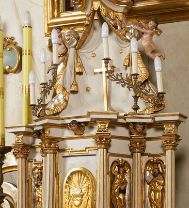 Zdjęcie nr 5: Tabernakulum architektoniczne, jednokondygnacyjne, pięcioosiowe. Osie wydzielone są sześcioma kanelowanymi kolumienkami na wysokich, profilowanych cokołach, z liściastymi kapitelami, podtrzymującymi profilowane belkowanie wyłamane na ich osiach. W polach pomiędzy kolumnami znajdują się nisze ujęte profilowaną ramą z uszakami. W czterech bocznych niszach, na trójdzielnych, profilowanych wspornikach stoją płaskorzeźbione postacie czterech ewangelistów na tle muszli palmetowych. Figurki są całopostaciowe, w ujęciu frontalnym, w złotych szatach, podtrzymują otwarte księgi, wskazując na tekst. Drzwiczki znajdują się na osi środkowej, zamknięte są półkoliście, płaskorzeźbione z przedstawieniem kielicha z koroną cierniową i z hostią w glorii promienistej. Kielich flankowany jest kłosami zbóż i winną latoroślą oraz winogronami. W partii belkowania płaskorzeźbiona dekoracja złożona z naprzemiennie ułożonych główek anielskich o złotych skrzydłach i płaskich rozetek. Tron wystawienia w formie baldachimu, z lambrekinem i chwostami, zwieńczony jest kulą z krzyżem, z podwieszonym z tyłu paludamentem podtrzymywanym przez parę puttów. Struktura polichromowana na biało, profile i detale pozłocone. 