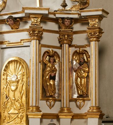 Zdjęcie nr 7: Tabernakulum architektoniczne, jednokondygnacyjne, pięcioosiowe. Osie wydzielone są sześcioma kanelowanymi kolumienkami na wysokich, profilowanych cokołach, z liściastymi kapitelami, podtrzymującymi profilowane belkowanie wyłamane na ich osiach. W polach pomiędzy kolumnami znajdują się nisze ujęte profilowaną ramą z uszakami. W czterech bocznych niszach, na trójdzielnych, profilowanych wspornikach stoją płaskorzeźbione postacie czterech ewangelistów na tle muszli palmetowych. Figurki są całopostaciowe, w ujęciu frontalnym, w złotych szatach, podtrzymują otwarte księgi, wskazując na tekst. Drzwiczki znajdują się na osi środkowej, zamknięte są półkoliście, płaskorzeźbione z przedstawieniem kielicha z koroną cierniową i z hostią w glorii promienistej. Kielich flankowany jest kłosami zbóż i winną latoroślą oraz winogronami. W partii belkowania płaskorzeźbiona dekoracja złożona z naprzemiennie ułożonych główek anielskich o złotych skrzydłach i płaskich rozetek. Tron wystawienia w formie baldachimu, z lambrekinem i chwostami, zwieńczony jest kulą z krzyżem, z podwieszonym z tyłu paludamentem podtrzymywanym przez parę puttów. Struktura polichromowana na biało, profile i detale pozłocone. 